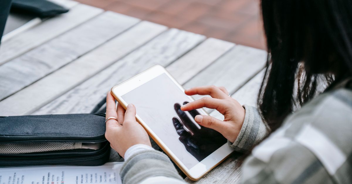 High angle of crop anonymous female student using tablet sitting at wooden table with papers in folder while doing homework in campus