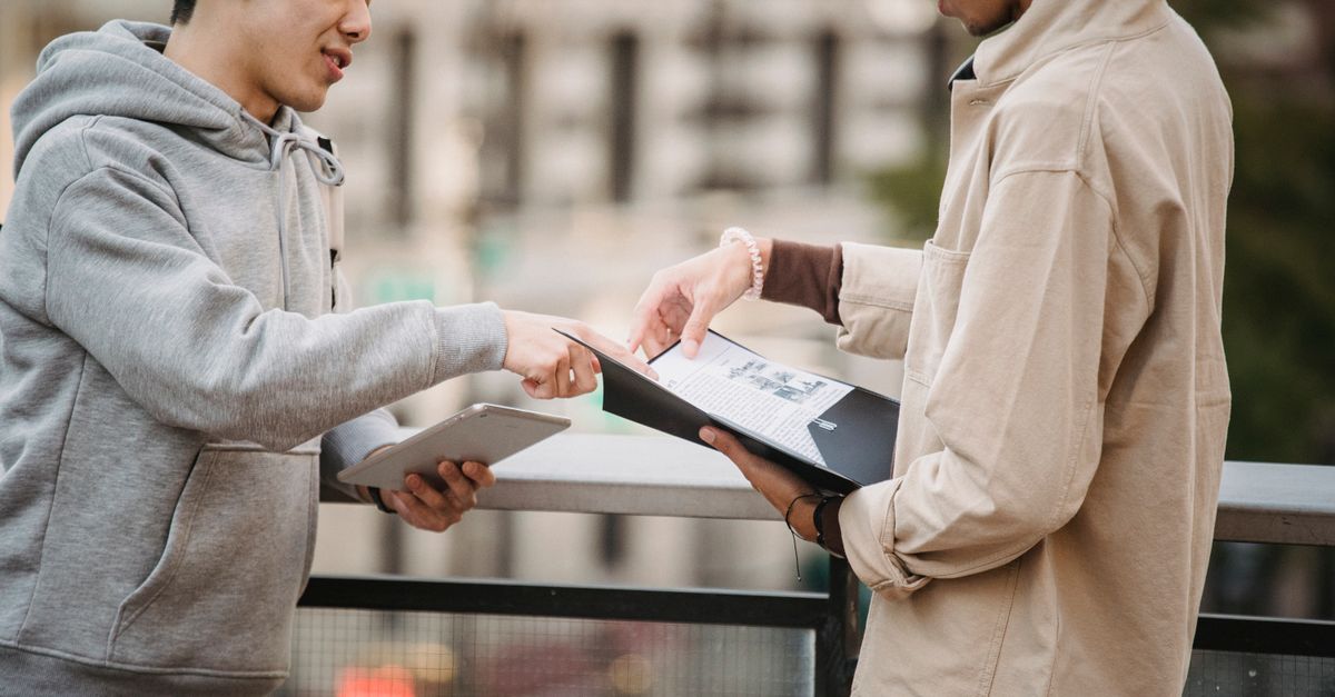 Side view of young diverse male students in casual clothes standing on street with tablet and papers in folder and speaking about home task